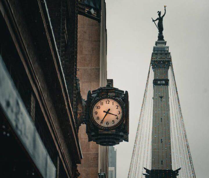 A clock on the side of a building with a statue in the background.